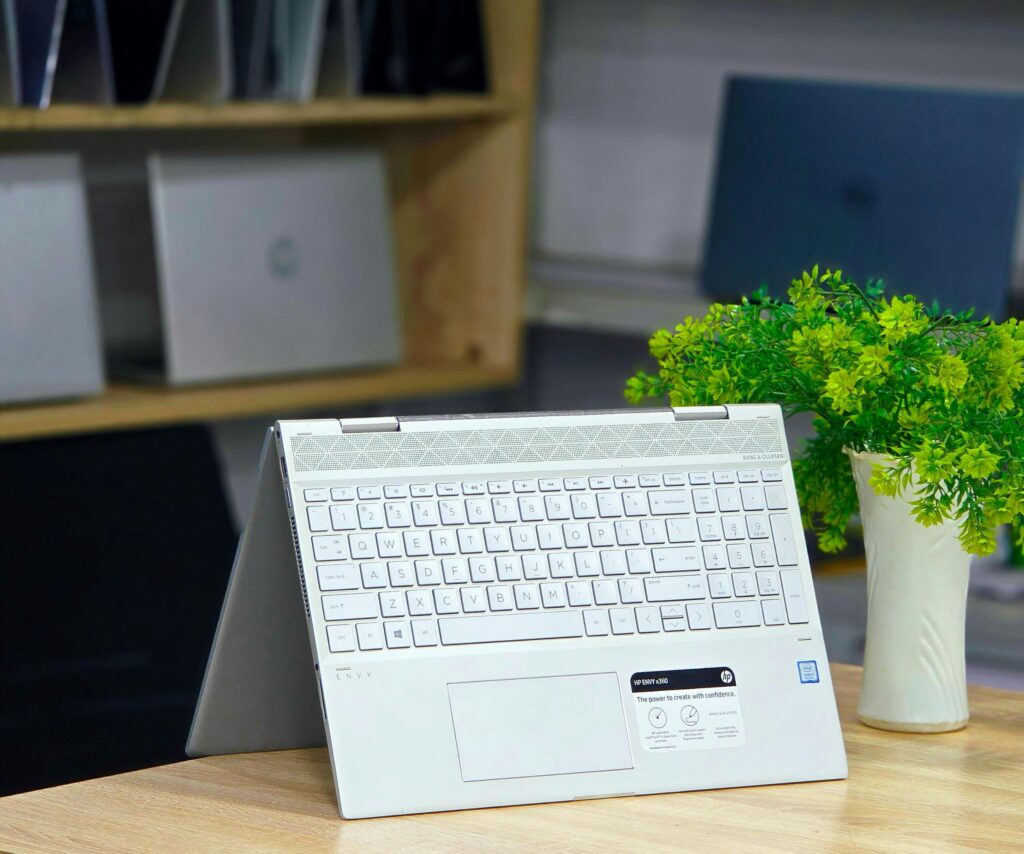 A modern convertible laptop on a wooden desk with a green plant in a bright office.